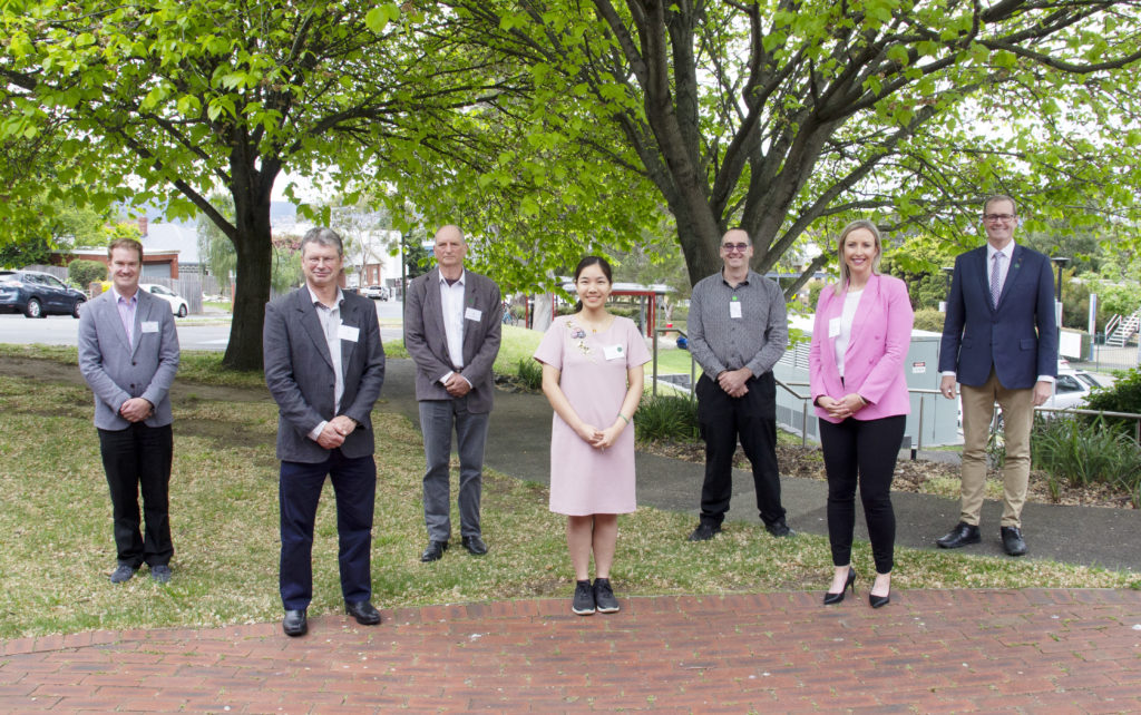 A mixed group of professionally dressed people stand socially distanced outside with nice trees in background.