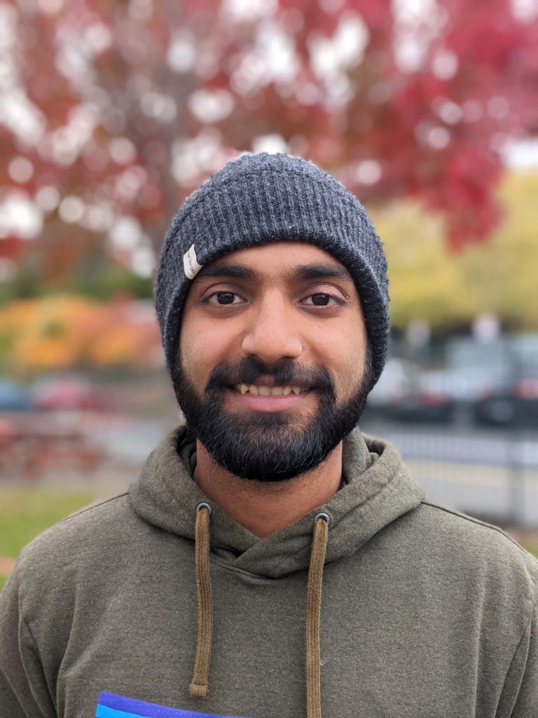 A man standing outside in autumn smiles. He is wearing a beanie, hoodie, and has a beard