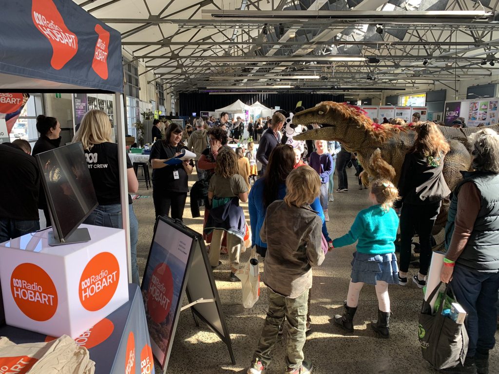 The ABC Hobart radio broadcast stall showing logos in a big round circle, and tons of visitors to the Festival of Bright Ideas in a crowd, various ages and clothing styles. It's in a warehouse with tall ceilings and conveys a noisy, fun atmosphere. 