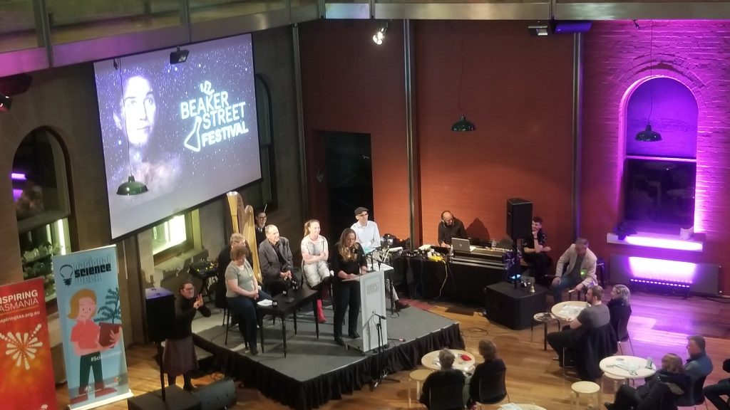 A mobile photo from above of speakers on a small stage at the Tasmanian Museum and Art Gallery open floor gallery, with some small tables with guests watching the speakers. There are 6 people on stage and a sign language translator to the side. Event signage includes Inspiring Tasmania, National Science Week, and Beaker Street Festival. Sound engineers sit to the right side. 