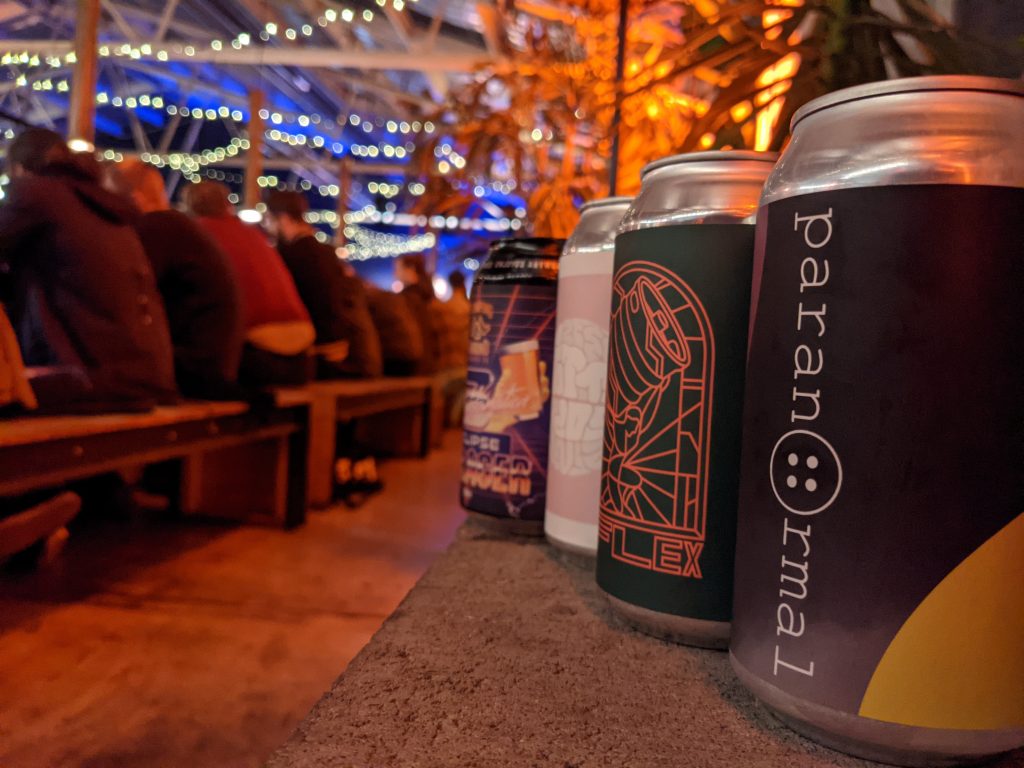 In the foreground is four different craft beer cans of varying designs. In the background is a long bench full of people with fairy lights all over the ceiling. It's a night time scene, contemporary pub atmosphere.